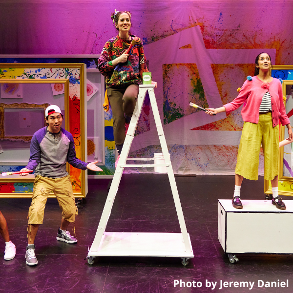 Picture frame and canvas backdrops with paint splatter and doodles. Three people holding brushes. Center person stands on a ladder. Right person stands on a rectangular platform.