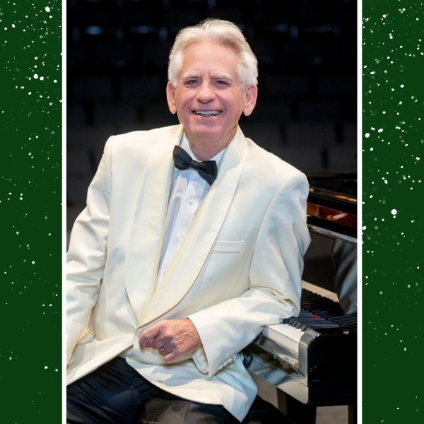 David Benoit sits next to a piano wearing a white suite and a black bowtie. He leans his elbow on the keys of the piano.