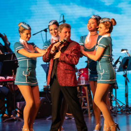 Male singer in a red suit surrounded by four stewardess in blue and white uniforms